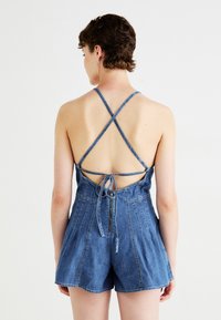 Denim playsuit met kruislussen en open rug, voorzien van een gestrikte sluiting en een ritssluiting aan de voorkant. Lichtblauwe kleur, korte lengte, gestructureerde stof.