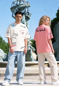 Jeune homme et femme portant des t-shirts graphiques avec des messages positifs, debout en plein air près d'une statue de globe et d'une fontaine sous un ciel clair.