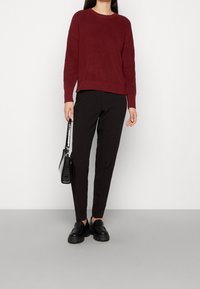 Burgundowy sweter z dzianiny z okrągłym dekoltem, w połączeniu z czarnymi dopasowanymi spodniami i czarnymi mokasynami. Trzyma małą czarn�ą torebkę na ramię.