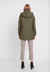 Giacca parka con cappuccio di colore verde oliva, caratterizzata da una texture trapuntata. Indossata sopra pantaloni a quadri, taglio cropped, e stivali neri alla caviglia. Vista da dietro.