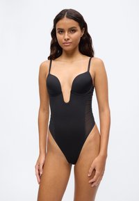 Frau mit langen dunklen Haaren, die einen schwarzen Badeanzug mit tiefem V-Ausschnitt und dünnen Schultergurten trägt, steht vor einem weißen Hintergrund.