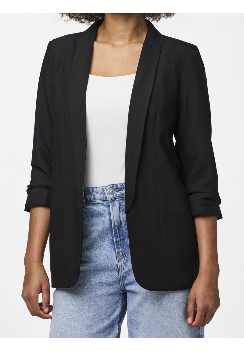 Femme portant un blazer noir sur un haut blanc avec un jean taille haute bleu clair, debout devant un fond blanc.