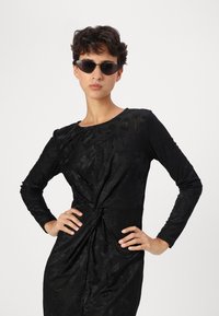 Vestido preto de mangas longas, padrão floral texturizado, detalhe de cintura anudada. Modelo usa óculos de sol estilo gato preto, com os braços cruzados contra um fundo branco.