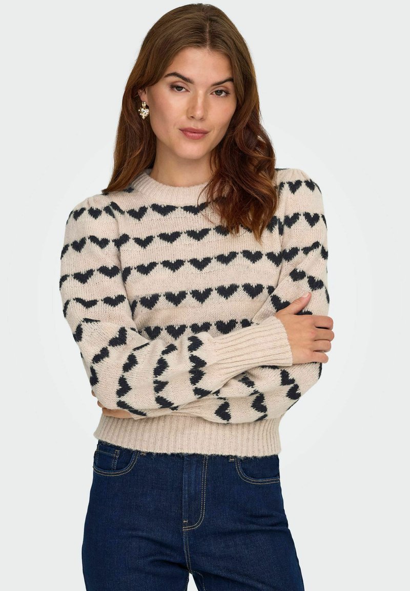 Suéter de punto en color crema con estampado de corazones negros, cuello redondeado y puños acanalados. Combinado con vaqueros oscuros de mezclilla.