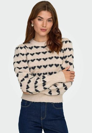 Pull en maille crème avec motif de cœur noir, col rond et poignets côtelés. Associé à un jean en denim foncé.