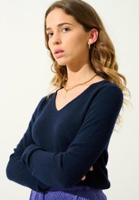 Navy v-ausschnitt Pullover aus weichem Stoff mit langen Ärmeln. Hat eine figurbetonte Form und wird mit einem strukturierten lila Unterteil kombiniert.