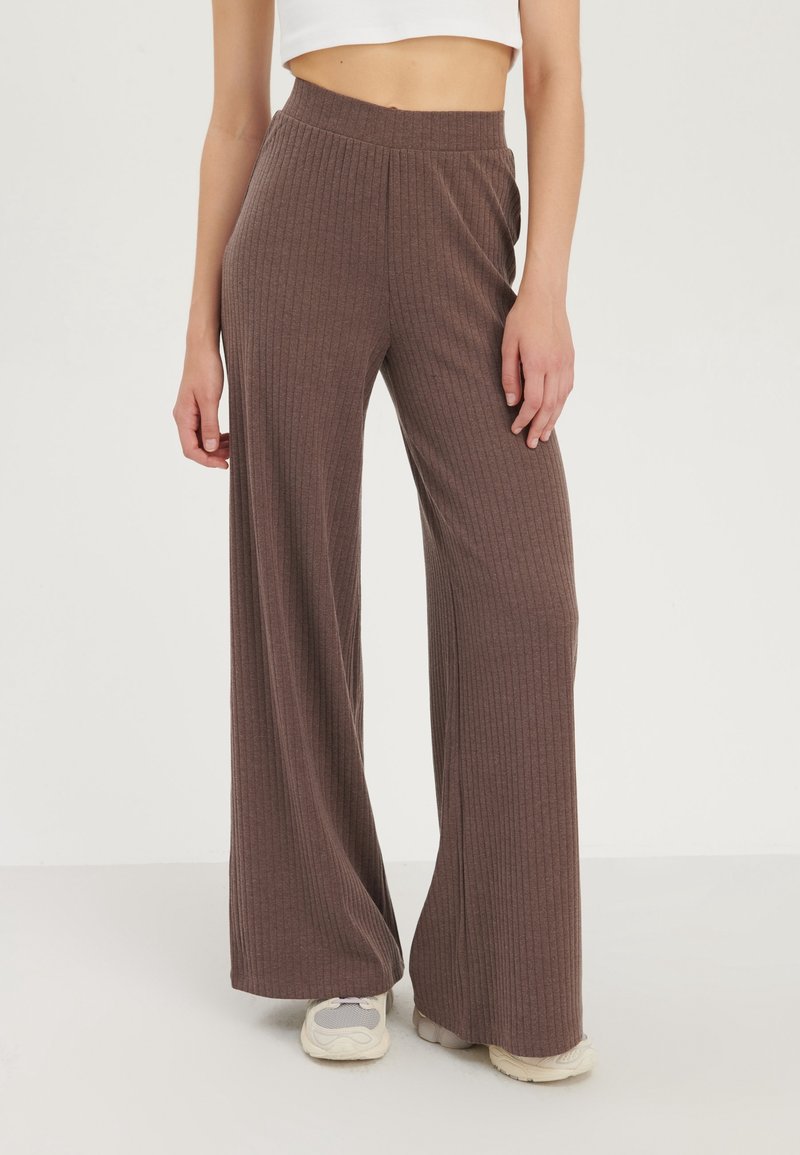 Brązowe szerokie spodnie z prążkowanym wzorem i wysokim elastycznym paskiem w talii, zaprojektowane z myślą o komforcie i elastyczności. W połączeniu z jasnoszarymi trampkami.