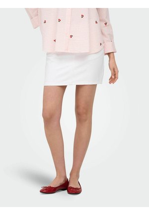 Personne portant une mini-jupe blanche, une chemise rayée rose et blanche avec petits motifs cerises, et des ballerines rouges, debout sur un fond uni.