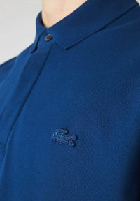 Polotröja i mörkblått, tillverkad av texturerat tyg, med en knappkrage och en liten broderad krokodil-logotyp på vänster bröst.