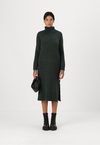 Abito verde lavorato a maglia con un alto colletto a coste, spacco laterale e maniche lunghe. Stivaletti neri alla caviglia e una piccola borsa nera completano il look.
