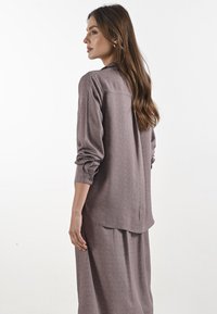 Blusa a maniche lunghe, dal taglio morbido, di colore malva con un motivo a pois testurizzato, con orlo leggermente curvo e dettaglio di pieghe sul retro.