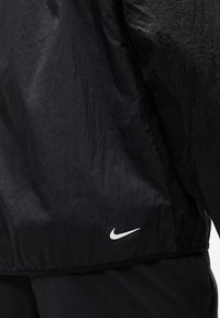 Μαύρο αθλητικό ύφασμα μπουφάν με διακριτική υφή και λευκό λογότυπο Nike Swoosh κοντά στο τελείωμα στο πίσω μέρος, φορεμένο πάνω από μαύρο παντελόνι.