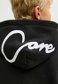 Sudadera negra con capucha que presenta un gran logo bordado en blanco en la parte posterior. La tela parece suave con una textura blanda alrededor del cuello y la capucha.