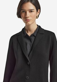 Schwarze Blazer aus strukturiertem Stoff, mit Ein-Knopf-Verschluss und Reverskragen, über einem dunkel gestreiften Hemd getragen. Das Model hat kurze Haare.