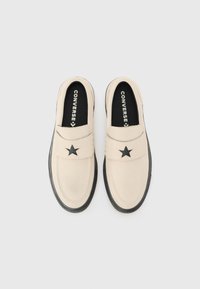 Béžové slip-on boty s černou hvězdou jako akcent na pásku, gumovou podešví a černým podšívkou s výrazně vytištěným nápisem 'CONVERSE'.