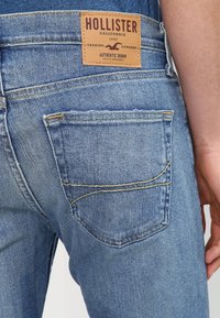 Jeans de mezclilla azul con una etiqueta de cuero marrón que dice "HOLLISTER", bolsillo trasero con detalle de costura y un diseño de pierna ajustada.