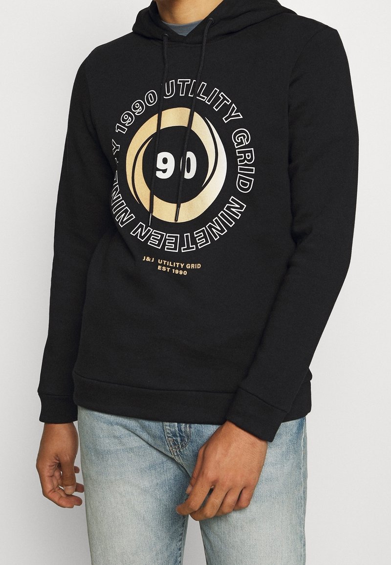 Felpa nera con grafica frontale che presenta un design circolare in oro e bianco, con la scritta "1990 UTILITY GRID" e il numero "90". Materiale misto cotone.