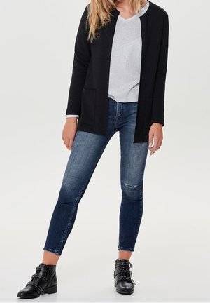 Femme portant un cardigan noir ouvert sur le devant, un haut gris clair à col en V, un jean skinny bleu et des bottines noires cloutées, debout sur un fond uni.