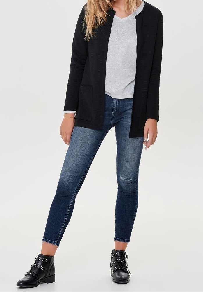 Femme portant un cardigan noir ouvert sur le devant, un haut gris clair à col en V, un jean skinny bleu et des bottines noires cloutées, debout sur un fond uni.