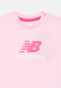 Light pink New Balance baby shirt with darker pink logo and white text, featuring a snap button on the shoulder neckline.