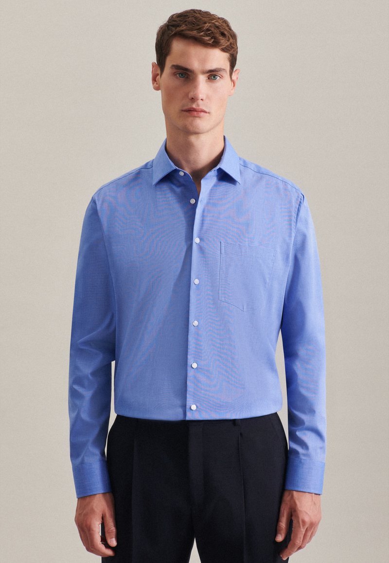 Camicia button-up azzurra con colletto a punta, taschino sulla sinistra del petto e maniche lunghe, abbinata a pantaloni scuri.