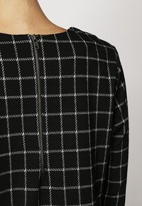 Černobílý kostkovaný oděv s vertikálním zipem, který vede dolů po horní části zad v oblasti ramene a krku osoby.