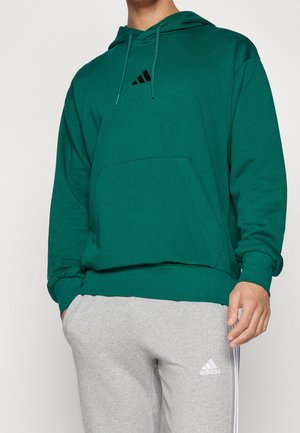 Homme portant un sweat à capuche vert avec un logo noir et un pantalon de survêtement Adidas gris clair avec des rayures blanches et un logo.