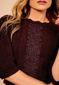 Žena v tmavě burgundy pleteném svetru s centrálním květinovým výšivkovým vzorem a polovičními rukávy.