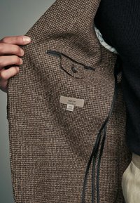 Braune Tweedjacke mit einem Innenfutter, einer Tasche, schwarzer Einfassung und einem Etikett mit der Aufschrift "NEXT REGULAR FIT". Die Textur scheint gewebt zu sein.