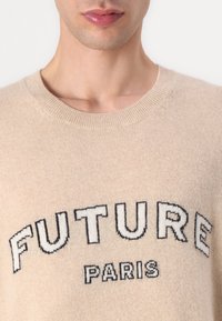 Beige strikkegenser med "FUTURE" og "PARIS" trykket i svart og hvitt. Rund hals, myk tekstur, og ribbestrikkede detaljer.