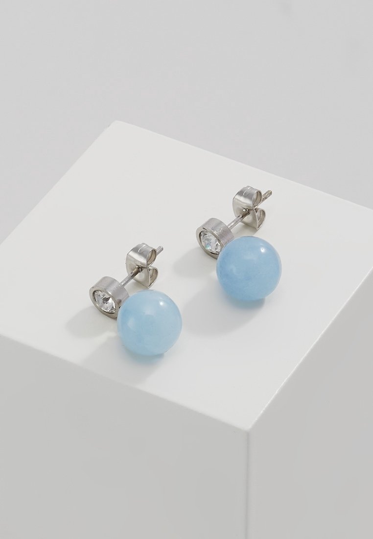 Boucles d'oreilles à clous en argent avec des pierres précieuses claires et des perles rondes bleu clair sur un piédestal blanc avec un fond gris.