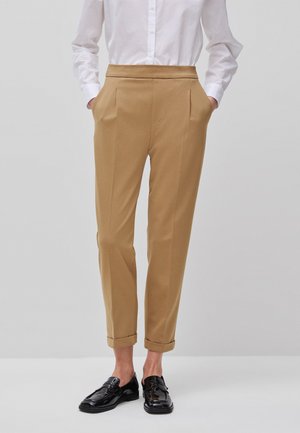 Osoba ubrana w beżowe, dopasowane spodnie ze spodenkami podwiniętymi na końcach, białą koszulę na guziki, z rękami w kieszeniach oraz czarne, błyszczące mokasyny na jednolitym tle.