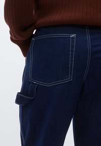 Pantalones de mezclilla azul oscuro con un bolsillo trasero visible con costuras blancas en contraste y un bolsillo lateral adherido a la parte superior del muslo.