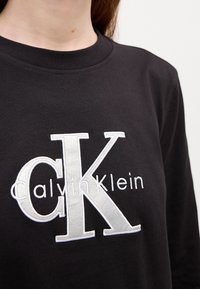 Juodas džemperis su gumuotu apykakle, priekyje su baltai siuvinėtu "CK" ir "Calvin Klein" užrašu. Minkšta audinio tekstūra.