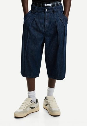 Denim-Shorts mit lockerer Passform, dunkelblauer Farbe, vorderen Falten und Seitentaschen. Getragen mit cremefarbenen Sneakers und weißen Socken.