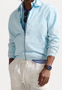 Ljusblå zip-up jacka med elastiska ärmslut och fåll, med sidofickor. Bärs över en randig skjorta, ihop med beiga byxor med snörning.