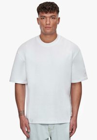 Hvid bomulds t-shirt med korte ærmer, afslappet pasform, rund halsudskæring og en diskret mærke-detail på venstre ærme. Glat tekstur.