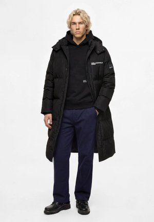 Junger Mann mit blondem Haar, der einen langen schwarzen Daunenmantel, einen schwarzen Hoodie, marineblaue Hose und schwarze Schuhe trägt und mit den Händen in den Taschen steht.