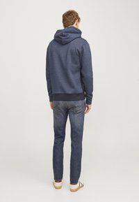 Sudadera azul marina con capucha, puños y dobladillo acanalados, combinada con jeans de mezclilla azules y zapatos de color claro, vista desde atrás.