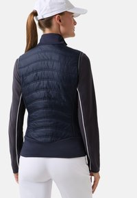 Marineblauw doorgestikt vest met contrasterende donkere mouwen en witte biezen. Voorzien van een hoge kraag en een strakke silhouet. Gestructureerde stofdetail op de achterkant.