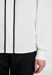 Valkoinen pitkähiha yhdistetty mustiin housuihin, joissa on kaksi pystysuoraa mustaa raitaa, käsi näyttäytyy sivussa.