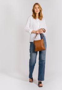 Brun läder crossbody-väska med en slät textur, rektangulär form och justerbar rem, matchad med en vit skjorta och blå jeans.