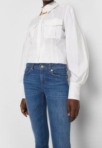 Vit långärmad skjorta med krage och två framfickor, i kombination med blå denimjeans. Har en kedjans accent vid halsringningen.