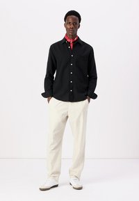 Camicia nera a maniche lunghe con bottoni e taschino sul petto, abbinata a pantaloni beige chiaro e un bandana rossa. Le scarpe sono bianche con dettagli marroni.