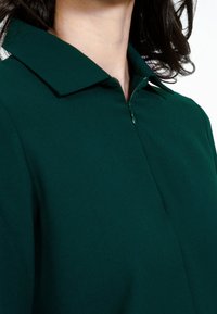 Tumši zaļā kleita ar apkakli, kas iekļauj priekšējo rāvējslēdzēju, izgatavota no gludas, nedaudz teksturētas auduma; tuvplānā uzmanība tiek pievērsta apkaklei un rāvējslēdzēja detaļām.