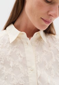 Light cream shirt with textured fringe detailing, a classic collar, and button closure; features a soft, sheer fabric.