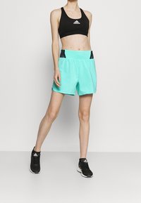 Persona vestida con un sujetador deportivo negro de Adidas, pantalones cortos turquesas con detalles negros y zapatillas negras de Adidas, de pie contra un fondo liso.