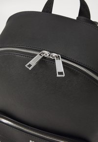 Sac à dos noir en tissu texturé, doté de deux fermetures éclair argentées et d'une poche inférieure zippée pour un rangement supplémentaire.