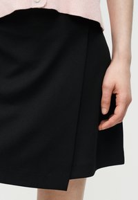 Femme portant une jupe noire longueur genou avec un panneau avant asymétrique et une manche de cardigan rose visible à la taille.