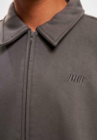Pelēks ar rāvējslēdzēju jaka ar salocītu apkakli, kurai kreisajā krūšu daļā ir izšūta "DEF" logo. Materiāls izskatās mīksts un gluds.